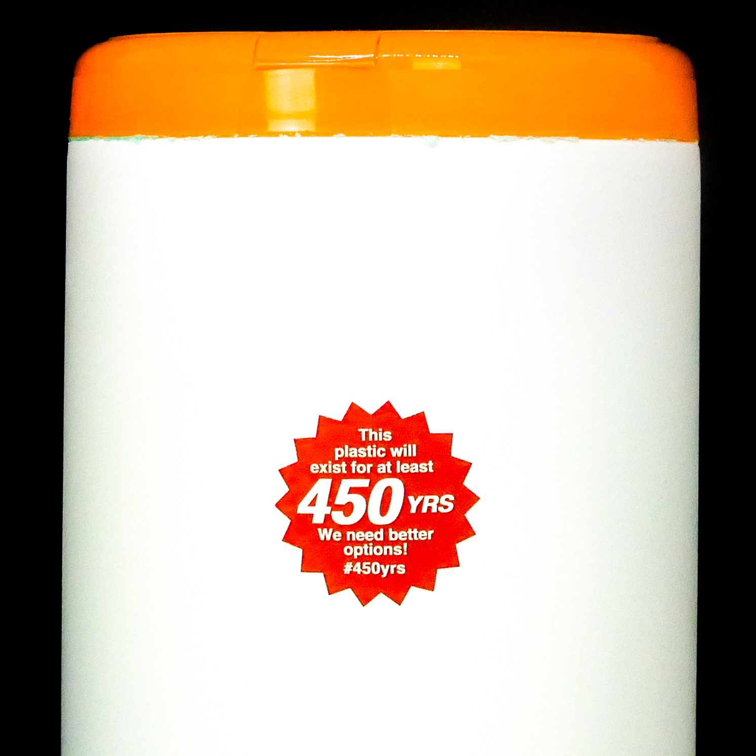 Thumbnail of Close-up photograph of a white, cylindrical shaped plastic container with an orange lid. Stuck on the container, is a red, starburst-shaped sticker that says, 'This plastic will exist for at least 450 YRS. We need better options! #450yrs.'